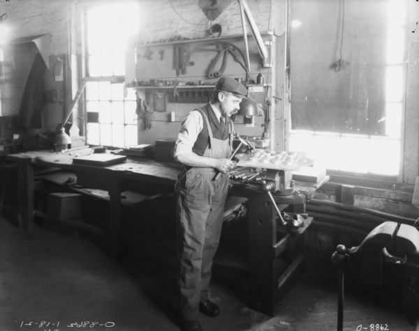 Male employee posing in finishing room near workbench. He is wearing overalls over a vest, shirt and necktie, as well as a hat. He is holding a hammer and a chisel above a piece of metal resting on top of a vise. Windows are along the wall above the workbench, and another vise is in the right foreground.