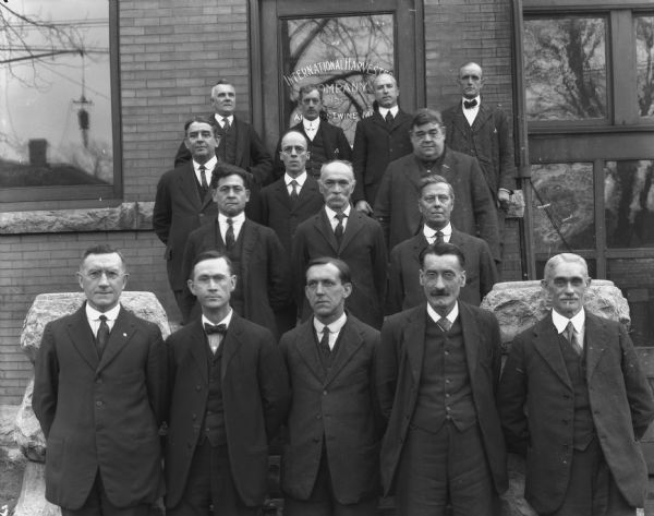 Fifteen male employees are posing in front of the entrance of a dealership. A sign on the glass of the front door reads: "International Harvester Company, Inc. Auburn Twin Mill."