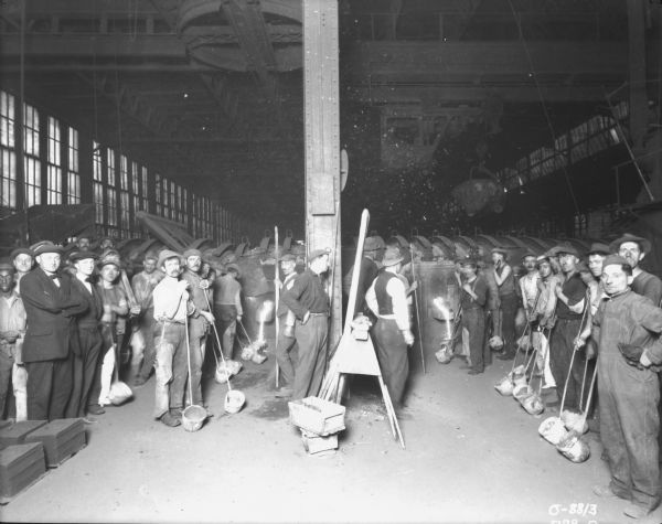 Group portrait of employees posing in foundry area, with many of the men holding work tools.