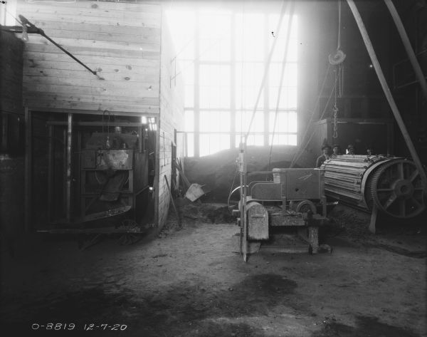 Interior view of the boiler/turbine room. Men are standing behind the turbine on the right, and a pile of coal is in front of a tall bank of windows in the center.