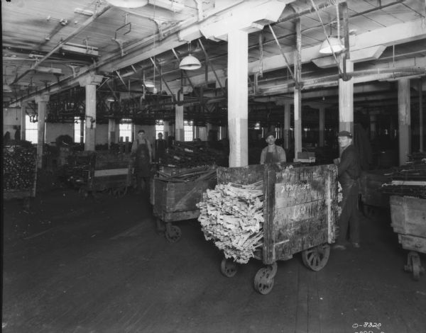 Men with are standing with carts loaded up with materials, probably metal parts. Windows are along the wall in the background.