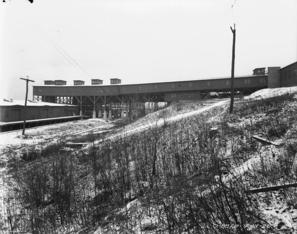 View across snow-covered hill towards a bridge near plant buildings. Railroad tracks and a building with a loading dock are on the left.