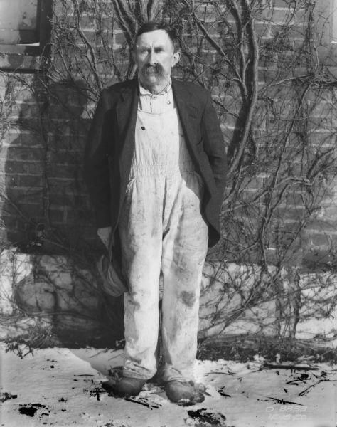 Portrait of a man wearing work clothes standing outdoors in front of a brick wall with vines growing up the sides. He is holding a hat in his right hand. Snow is on the ground.