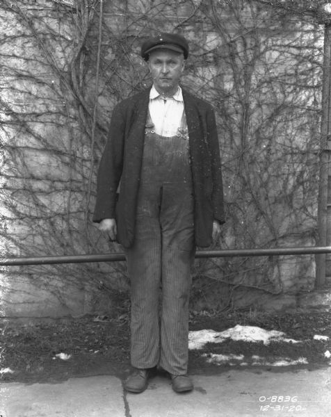 Portrait of a man wearing work clothes standing outdoors in front of a brick wall with vines growing up the sides. Some snow is on the ground.
