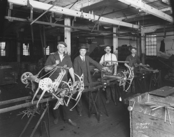 Four men are posing together with Binder parts. The man on the left is holding a hammer and is wearing work clothes. The man in the center is wearing a suit and hat, and the two men on the right are wearing work clothes.