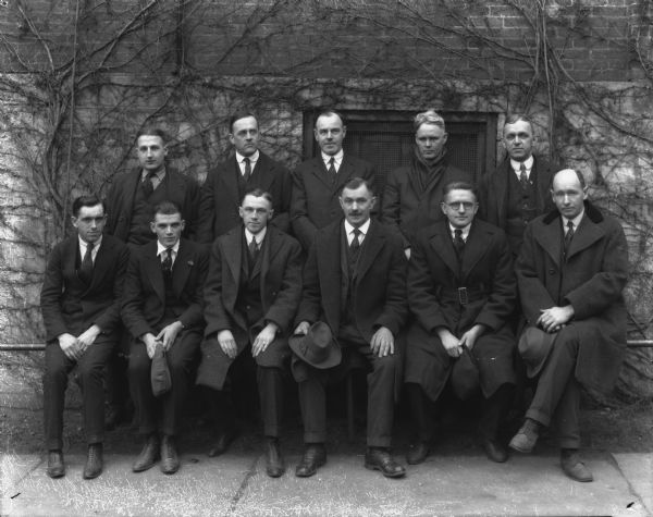 Group portrait of eleven men posing on and behind a railing in front of the brick wall of a factory building.