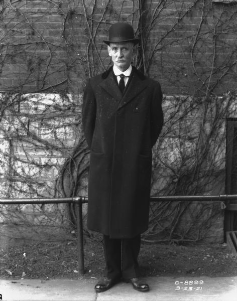 Full-length portrait of a man standing on a sidewalk in front of a railing along a brick wall of a factory building. He is wearing a hat and a three-quarter length coat over a suit.
