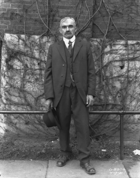 Full-length portrait of a man wearing a suit and holding a hat standing on a sidewalk in front of a railing along a brick wall of a factory building.