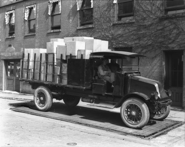View towards two men sitting in a truck loaded with boxes parked along the side of a brick building. The truck is on a wooden platform that may be a scale.