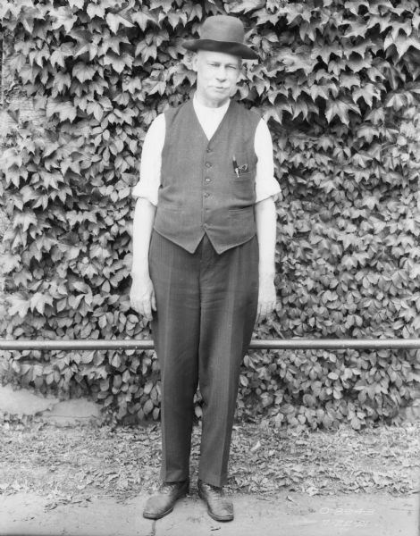Full-length portrait of a man standing on a sidewalk in front of a railing along a brick wall of a factory building. He is wearing a hat, and a vest over a shirt and trousers.