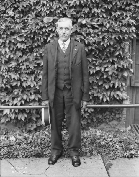 Full-length portrait of a man standing on a sidewalk in front of a railing along a brick wall of a factory building. He is wearing a suit with a vest and necktie, and is holding a straw boater in his right hand.