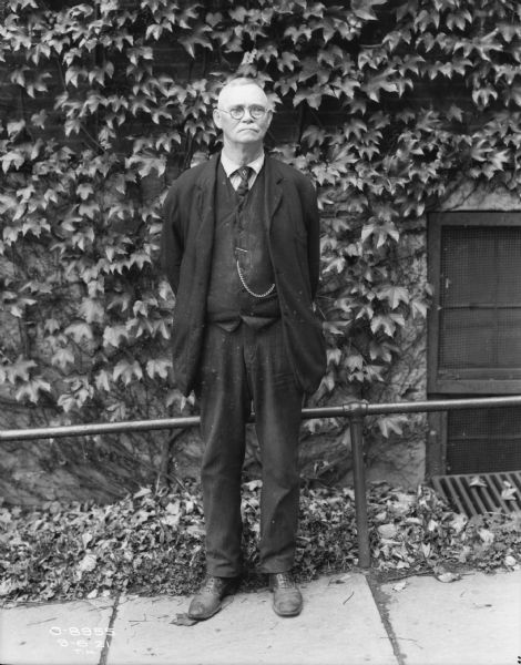 Full-length portrait of a man standing on a sidewalk in front of a railing along a brick wall of a factory building. He is wearing a suit and eyeglasses.