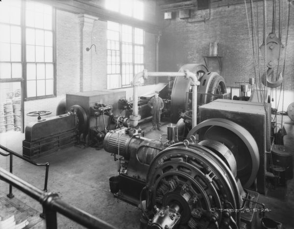 View over railing looking down over generators inside the plant. A man is standing in the center among the machinery. Tall windows are along the brick factory wall on the left.