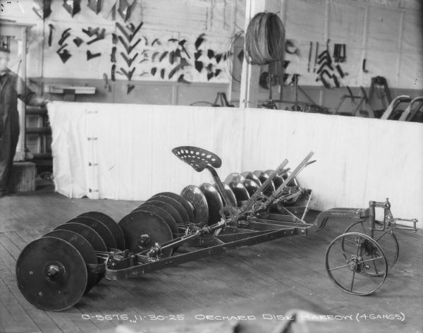 Orchard Disk Harrow (4 Gangs) set up on factory floor with a low backdrop behind. A man is standing on the far left holding up one end of the backdrop. Parts are hanging on the wall in the background.