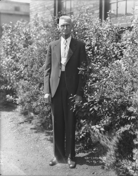 Full-length portrait of a man wearing a business suit and eyeglasses standing near bushes alongside a brick building.