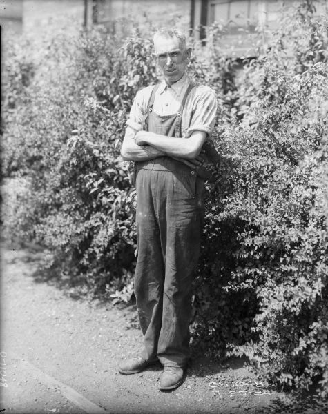 Full-length portrait of a man wearing overalls with his arms across his chest standing near bushes alongside a brick building.