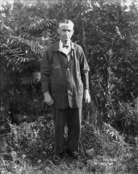 Full-length portrait of a man wearing a lab coat over a shirt, bowtie and pants, standing outdoors near in tall grass near trees.