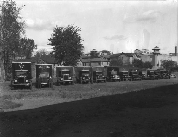 Two men are standing in front of a row of 13 vehicles of the Red Star Express Lines fleet. In the background are houses and dwellings.