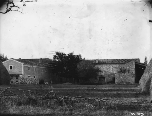View of farmyard, with stone farm buildings, with large haystacks on the left and right. Location: France.