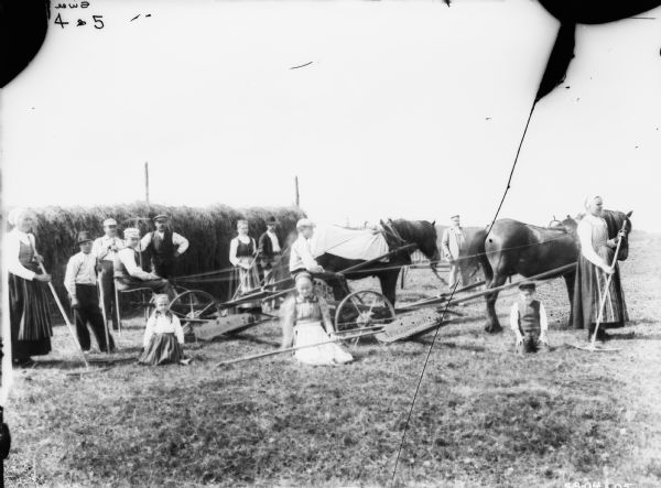 Large group of men, women and children posing in a field. Two men are on horse-drawn McCormick mowers. The rest of the group is holding agricultural implements. A large stack of hay is in the background. Location: France.