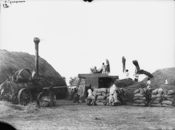 Men are using a tractor to belt-drive a thresher in a field. Women are standing near a ladder in the center. A man wearing a hat, long coat, and holding an umbrella is standing near piles of sacks on the right. Large haystacks are on the left and right. Location: Hungary.