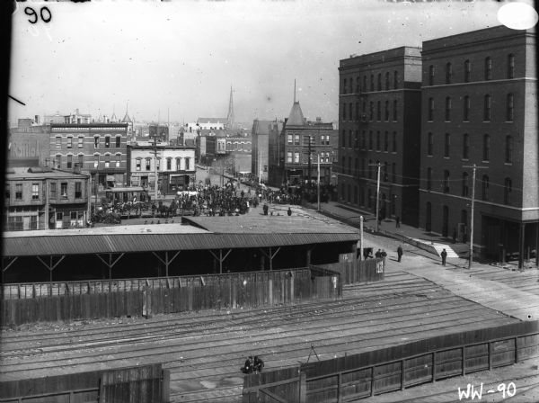 Elevated view over railroad tracks towards a crowd of people along a commercial street near horse-drawn vehicles and a street railroad car.