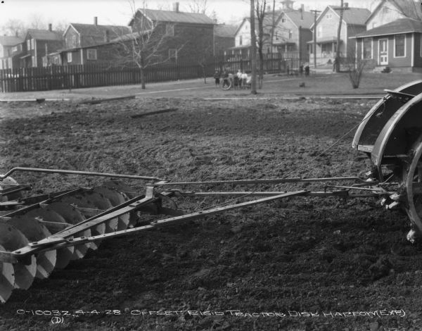 Disk Harrow drawn by tractor sitting in a field, with a view of neighborhood beyond. Children are watching in the background. A fence and houses are behind them. Test plot?