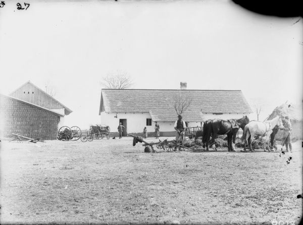 Family posing with implements for farming. Men are standing on the right with a team of two horses and agricultural equipment. In the background a man, young girl, and a woman holding a child in her arms are standing in front of a building. Location: Hungary.