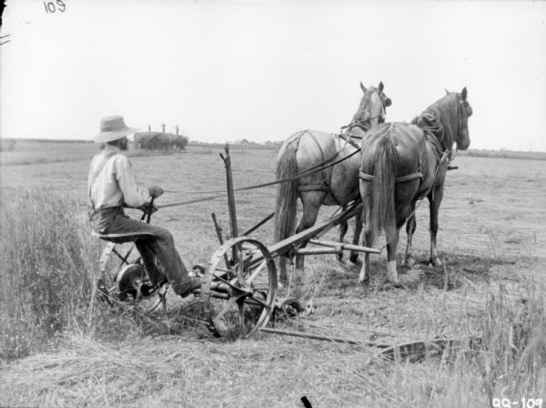 Rear view of a man on a horse-drawn Mower in a field. In the background three people are standing on top of a large haystack.