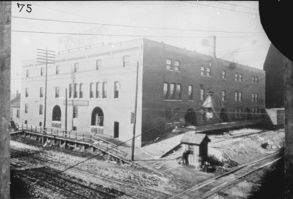 A man is standing near a small building at bottom right. A loading dock is along the large brick building on the left and right. Railroad tracks are in the right foreground.