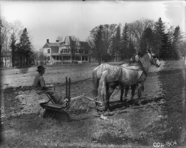 Right side view of a man riding a horse-drawn Disk Harrow in a field. There are buildings in the background, including a large house with a porch and balcony. 