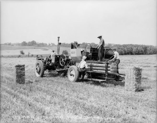 Four men are using a Farmall tractor with a pickup baler in a field. Farm buildings are on a hill in the distance.