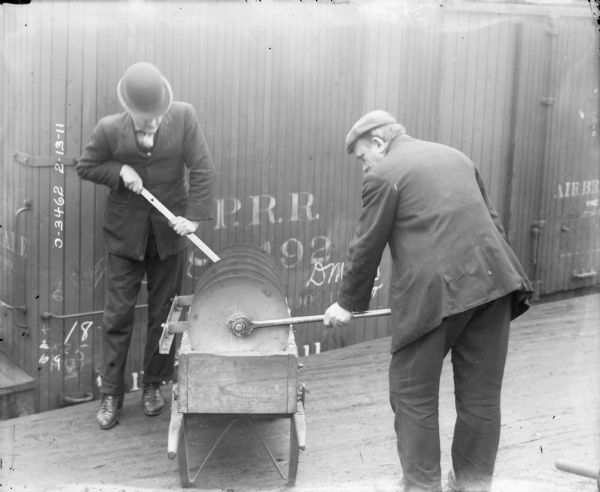 Two men adjusting gangs on a Disk Harrow part that is resting on a cart. There is a railroad car behind them.