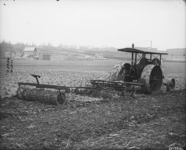 Three-quarter view from right rear towards a  Disk Harrow attached to a tractor in a field. There are farm and factory buildings in the distance.