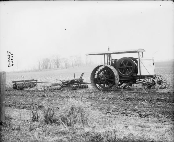 View through a fence of the right side of a tractor pulling a Disk Harrow in a field.