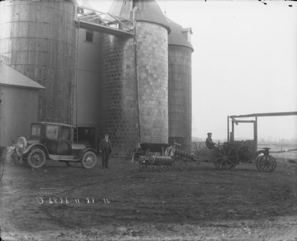A man is driving a tractor pulling Disk Harrows in a barnyard. A man is standing in the center, and an automobile is parked on the left near farm buildings and silos.