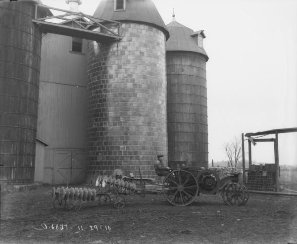 Right side profile view of a farmer posing driving a tractor pulling a Disk Harrow. Silos and a large barn are behind the farmer.