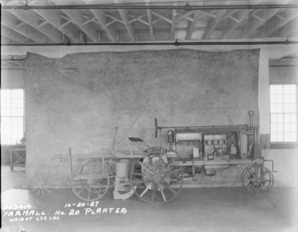 Right side, profile view of a Farmall Drawn Planter, No. 20, 632 lbs., parked indoors on factory floor. A backdrop is hanging from the ceiling.