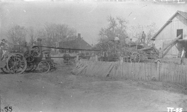 A tractor on the left, probably steam powered, is belt-driving an Ensilage Cutter. Men are working on and around a wagon near a barn.
