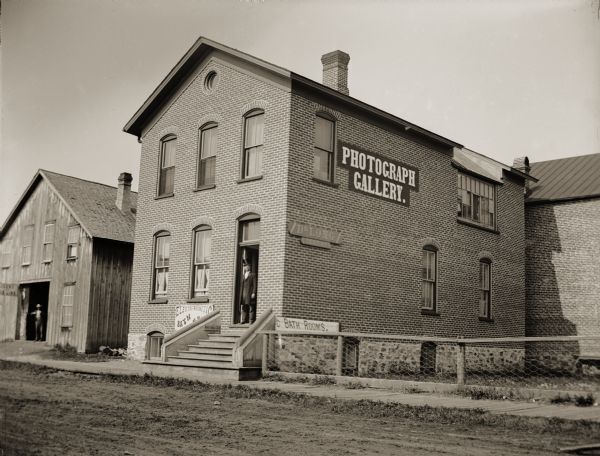 View across street towards the building that housed the gallery of Van Schaick and the dental offices of Edward F. Long. Over the years, the building also accommodated a newspaper office and a telephone company. There is an unidentified man posing standing in the doorway wearing a top hat. The Livery and U.S. Line in the building on the left.