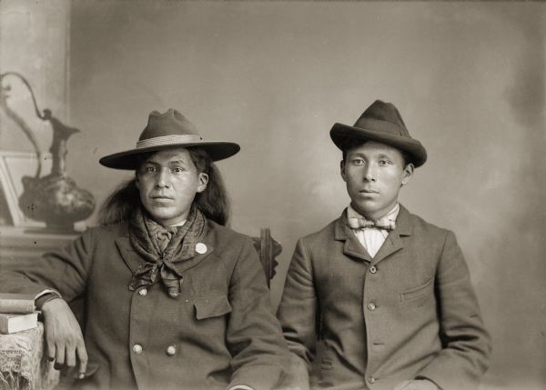 Waist-up studio portrait in front of a painted backdrop of Sam Carley Blowsnake (HoChunkHaTeKah), wearing a hat and sporting long hair, is sitting next to Martin Green (Snake) (KeeMeeNunkKah). Long hair was not a common hairstyle for Ho-Chunk men during this time period, but was adopted by some who performed in Wild West shows.