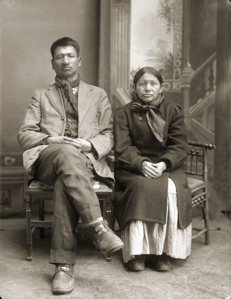 Full-length studio portrait of Thomas Thunder (HoonkHaGaKah) (Son of [WaConChaKah] John Thunder aka Dr. Thunder and [WeHonPeKaw] Lucy Bear, Thunder), and his wife, Addie Littlesoldier, Lewis, Thunder (KzunchJeKayRaWinKah) (Daughter of [NoGinKaw] Little Soldier and Mrs. ChoNeWeKaw). He is wearing a scarf around his neck, with a suit jacket, vest, and trousers. She is wearing a coat over a dress. In the background is a painted backdrop.