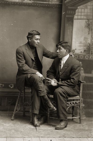 Full-length studio portrait of two Ho-Chunk men posing sitting in front of a painted backdrop. George Lonetree (HoonchXeDaGah), left, is sitting next to Robert Greengrass (HoNutchNaCooMeeKah). Both men are wearing suit jackets and trousers.