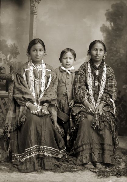 Full-length studio portrait of two Ho-Chunk women posing sitting in front of a painted backdrop near a prop wooden fence. The women are wearing earrings, rings, and bead necklaces. A young Ho-Chunk boy is posing standing between the two women. They are identified from left to right as, Margaret Whitedeer Littlesam (NauChoPinWinKah), Lee Dick Littlesam (HaNotchANaCooNeeKah), and Emma Stacy Whitedeer Lowery (HompInWinKah).