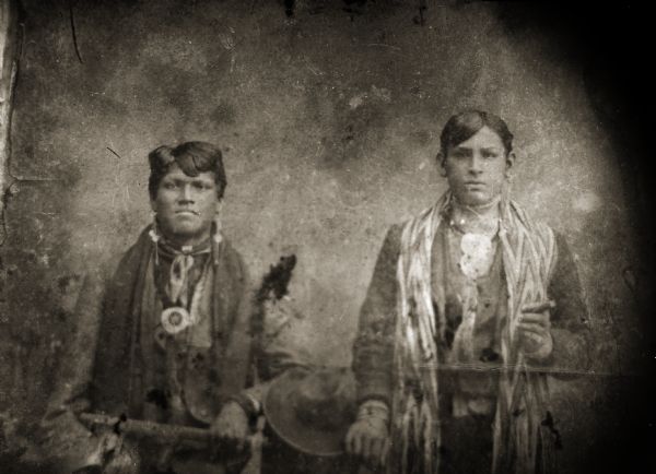 Copy photograph of a studio portrait of two young Ho-Chunk men posing standing and wearing regalia, including a finger-braided shawl on the man on the right. The man on the left is probably holding a metal tomahawk pipe. Identified as Henry Thunder (WahQuaHoPinKah), left, and George Funmaker Sr. (WojhTchawHeRayKah) holding a cigar on the right.