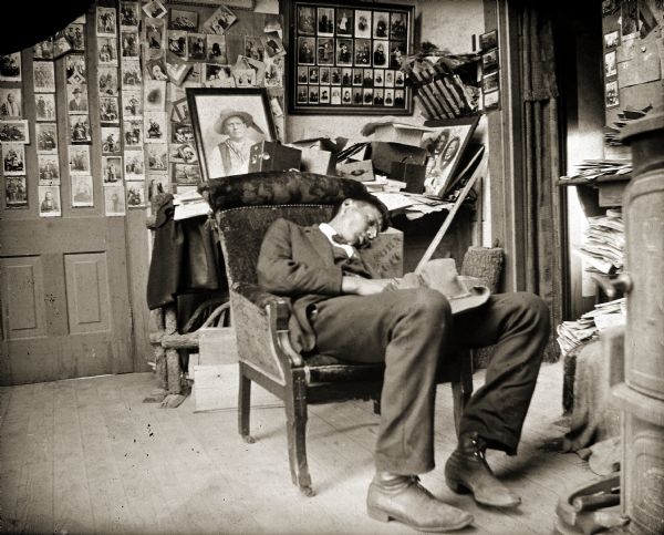 A man sleeping in a chair, possibly Thomas Thunder (HoonkHaGaKah). On the back wall of Van Schaick’s studio are photographs he made available for purchase.