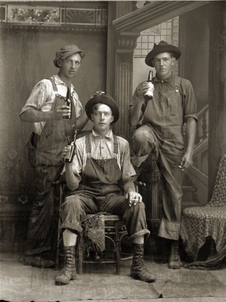 Full-length studio portrait in front of a painted backdrop of three unidentified white men dressed in bib overalls. One man is sitting in the center between the two other men, who are standing. The men are holding cigarettes and beer bottles.