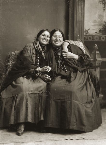 Full-length studio portrait in front of a painted backdrop of two smiling Ho-Chunk (Winnebago) women identified as Clara St. Cyr, a Nebraska Winnebago, left, and Lucy Davis (NukZeeKah), the sister of John Davis. They are holding a bottle between them that has been retouched.