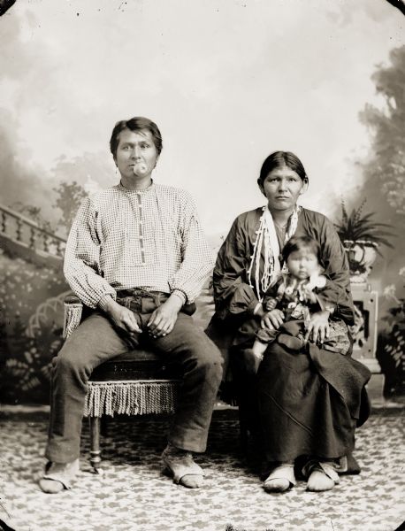 Studio portrait in front of a painted backdrop of an unidentified man sitting with his family, and holding an apple in his mouth. The Ho-Chunk woman is posing sitting on the right wearing several necklaces, and a pair moccasins. She is holding a small child on her lap.