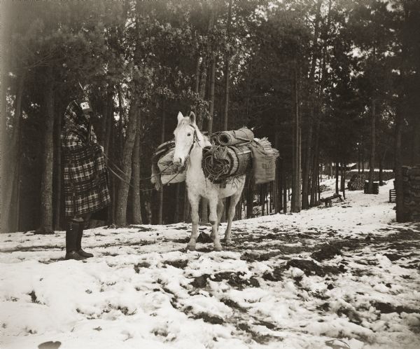 David Goodvillage (WauHeTonChoEKah), standing with his horse who is loaded with supplies, including cattail mats, snowshoes and canvases, for a hunting and trapping trip at Marshland near the Mississippi River. There is a building and a number of large stacks of fuelwood in the background on the right.

